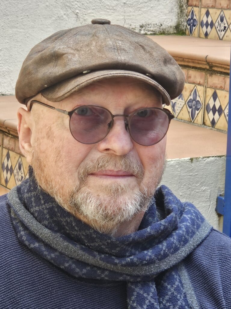 Mark Bertrand wearing a flat cap, tinted glasses, and scarf, seated on outdoor steps.